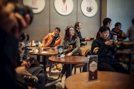 A group of people sitting around tables in a cafe, each person either holding a camera or a smartphone. The setting appears cozy, with warm lighting and soft brown tones. Some people are focused on their devices, while others seem relaxed, engaging with their surroundings.