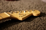 A close-up of the headstock of a guitar, with tuning pegs and a logo visible. The guitar neck is resting on a textured surface.