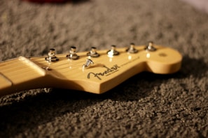 A close-up of the headstock of a guitar, with tuning pegs and a logo visible. The guitar neck is resting on a textured surface.