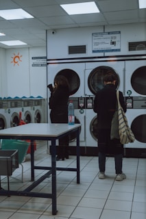 A laundromat interior with two people standing near a row of front-loading washing machines. One person is facing away, holding a striped bag and wearing casual shoes. The other person is also facing the machines, with a bottle placed on a table in the foreground. The room is brightly lit with fluorescent lights, and there are instructions displayed above the machines.