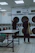 A laundromat interior with two people standing near a row of front-loading washing machines. One person is facing away, holding a striped bag and wearing casual shoes. The other person is also facing the machines, with a bottle placed on a table in the foreground. The room is brightly lit with fluorescent lights, and there are instructions displayed above the machines.
