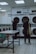 A laundromat interior with two people standing near a row of front-loading washing machines. One person is facing away, holding a striped bag and wearing casual shoes. The other person is also facing the machines, with a bottle placed on a table in the foreground. The room is brightly lit with fluorescent lights, and there are instructions displayed above the machines.