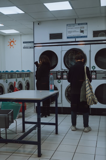 A laundromat interior with two people standing near a row of front-loading washing machines. One person is facing away, holding a striped bag and wearing casual shoes. The other person is also facing the machines, with a bottle placed on a table in the foreground. The room is brightly lit with fluorescent lights, and there are instructions displayed above the machines.