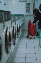 A row of washing machines lines the left side of a tiled-floor laundromat. A person is placing items into or removing items from one of the machines. There is a red shopping trolley beside them.