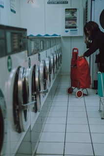 A row of washing machines lines the left side of a tiled-floor laundromat. A person is placing items into or removing items from one of the machines. There is a red shopping trolley beside them.