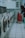 A row of washing machines lines the left side of a tiled-floor laundromat. A person is placing items into or removing items from one of the machines. There is a red shopping trolley beside them.