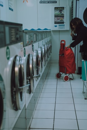 A row of washing machines lines the left side of a tiled-floor laundromat. A person is placing items into or removing items from one of the machines. There is a red shopping trolley beside them.