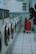 A row of washing machines lines the left side of a tiled-floor laundromat. A person is placing items into or removing items from one of the machines. There is a red shopping trolley beside them.