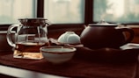 A serene tea setting featuring a glass pitcher with tea, a dark ceramic teapot, and a few tea cups on a textured surface. The background consists of large windows, allowing soft, natural light to fill the space, creating an inviting and calming atmosphere.
