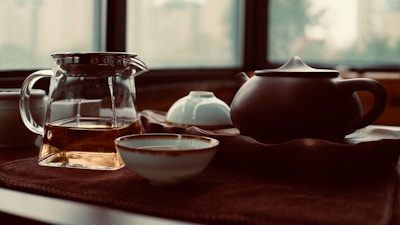 A serene tea setting featuring a glass pitcher with tea, a dark ceramic teapot, and a few tea cups on a textured surface. The background consists of large windows, allowing soft, natural light to fill the space, creating an inviting and calming atmosphere.