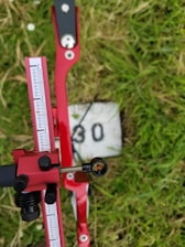 A red adjustable archery sight with measurement markings is in focus, set against a grassy background with a painted number 30 on a white block. The sight includes a small circular aiming aperture.
