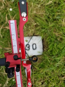 A red adjustable archery sight with measurement markings is in focus, set against a grassy background with a painted number 30 on a white block. The sight includes a small circular aiming aperture.