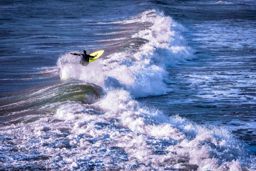 A surfer in a wetsuit skillfully rides a wave, balancing on a bright yellow surfboard as the ocean's waves crest and crash around them. The water appears lively and turbulent, capturing the dynamic motion of the sport.
