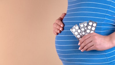 A person holding three blister packs of white pills against their pregnant belly. The individual is wearing a blue and white striped shirt, and the background is a plain beige color.