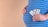 A person holding three blister packs of white pills against their pregnant belly. The individual is wearing a blue and white striped shirt, and the background is a plain beige color.