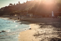 The image depicts a serene beach scene during sunset with a soft golden light illuminating the sandy shore. A few people are walking along the water's edge, and waves gently lap the sand. In the background, there are lush trees and beach amenities, including umbrellas and lounge chairs.