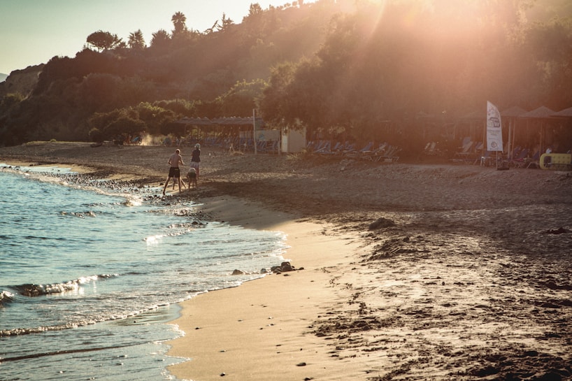 The image depicts a serene beach scene during sunset with a soft golden light illuminating the sandy shore. A few people are walking along the water's edge, and waves gently lap the sand. In the background, there are lush trees and beach amenities, including umbrellas and lounge chairs.