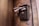 A textured, vintage-style mailbox is mounted on a stone wall. It is overflowing with newspapers and mail. Below the mailbox, there is a modern intercom with a metallic finish. The wall surface has a rustic appearance, with visible marks and discoloration.