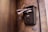 A textured, vintage-style mailbox is mounted on a stone wall. It is overflowing with newspapers and mail. Below the mailbox, there is a modern intercom with a metallic finish. The wall surface has a rustic appearance, with visible marks and discoloration.