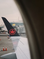 A view from an airplane window showing the wing with a logo featuring a red maple leaf on a black tail. The window frames the view, and in the background, there are airport buildings, a boarding gate, and several orange traffic cones on the tarmac.