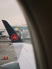 A view from an airplane window showing the wing with a logo featuring a red maple leaf on a black tail. The window frames the view, and in the background, there are airport buildings, a boarding gate, and several orange traffic cones on the tarmac.