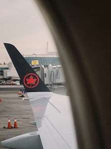 A view from an airplane window showing the wing with a logo featuring a red maple leaf on a black tail. The window frames the view, and in the background, there are airport buildings, a boarding gate, and several orange traffic cones on the tarmac.