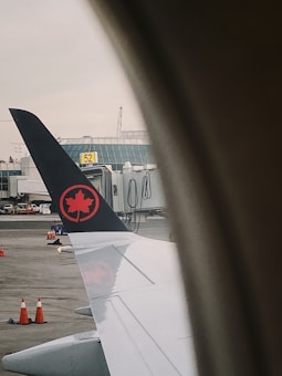 A view from an airplane window showing the wing with a logo featuring a red maple leaf on a black tail. The window frames the view, and in the background, there are airport buildings, a boarding gate, and several orange traffic cones on the tarmac.