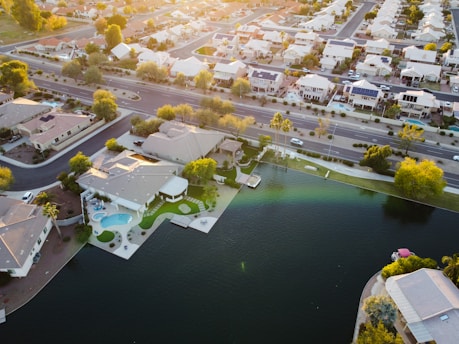 An aerial view of a suburban neighborhood with rows of houses lining the streets. The scene includes a small lake with a few adjacent houses featuring backyards and swimming pools. Several cars are parked on the roadway, and the trees are interspersed throughout the area, suggesting a well-planned residential environment.