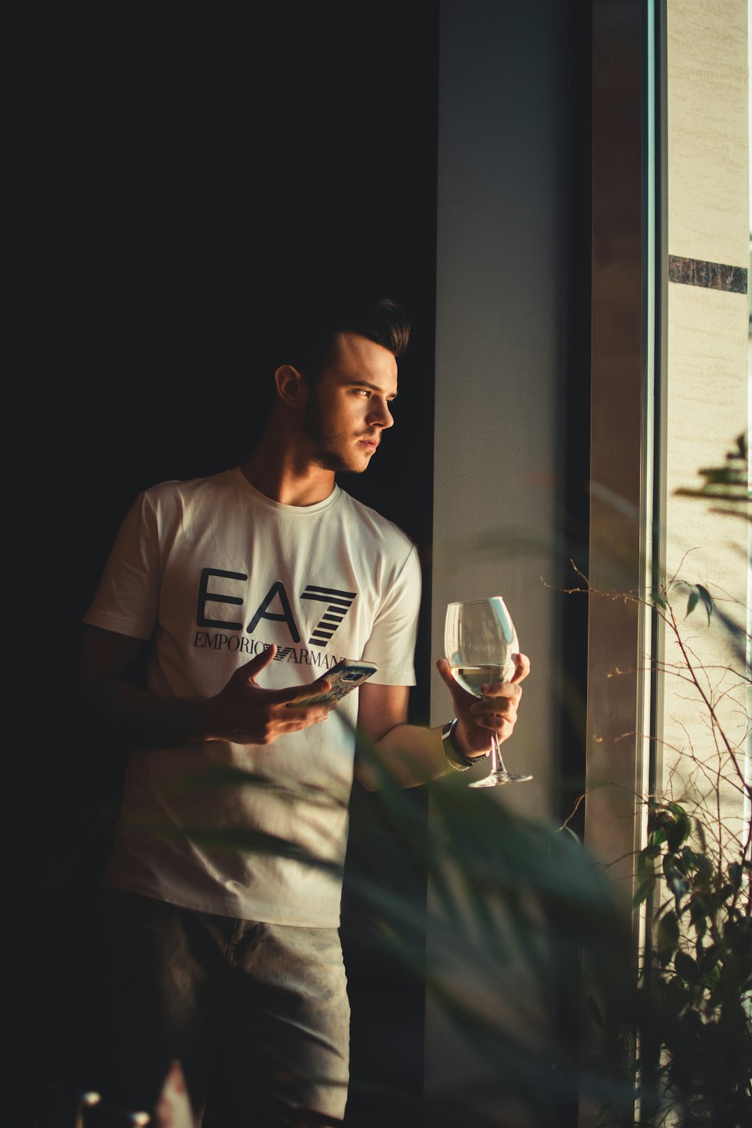 man in white EA7 t-shirt holding wine glass beside window