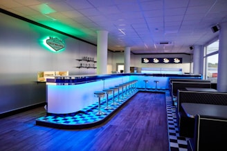 A modern, retro-style diner with a neon-lit bar and chrome stools, featuring a checkerboard floor pattern. The interior includes sleek black booths and a welcoming atmosphere accentuated by soft blue lighting. A sign on the wall reads 'Circuit Mallorca' alongside vintage decor elements.