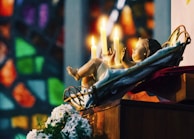 A religious scene features a figure of a child lying in a cradle, surrounded by glowing candles. The soft flickering light illuminates the scene, creating a warm and serene atmosphere. In the background, colorful stained-glass windows add a vibrant array of colors to the composition. White flowers are placed nearby, enhancing the peaceful setting.