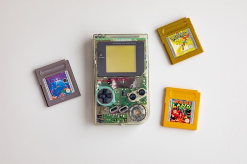A vintage Nintendo Game Boy with a clear casing is surrounded by three game cartridges. The cartridges include Tetris in a gray casing, Pokémon in a yellow casing, and Donkey Kong Land also in a yellow casing. The clear casing of the Game Boy reveals its internal circuitry.