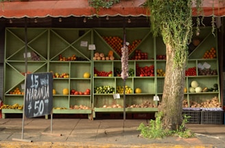 A rustic fruit and vegetable stand with wooden shelves displaying various produce items including oranges, apples, tomatoes, and pumpkins. A chalkboard sign advertises oranges at a price. The stand is partially covered by a tree with hanging vines adding a natural touch to the scene.