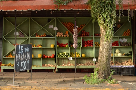 A rustic fruit and vegetable stand with wooden shelves displaying various produce items including oranges, apples, tomatoes, and pumpkins. A chalkboard sign advertises oranges at a price. The stand is partially covered by a tree with hanging vines adding a natural touch to the scene.