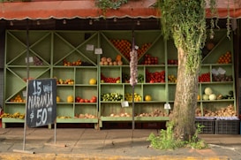 A rustic fruit and vegetable stand with wooden shelves displaying various produce items including oranges, apples, tomatoes, and pumpkins. A chalkboard sign advertises oranges at a price. The stand is partially covered by a tree with hanging vines adding a natural touch to the scene.