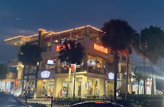 A two-story building illuminated at night showcases a Hooters restaurant on the upper level and a Ron Jon Surf Shop on the lower level. The structure is surrounded by palm trees, and bright lights highlight the architectural details. Several people can be seen near the entrances, with festive decorations including a wreath adding to the nighttime atmosphere.