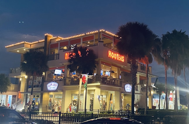 A two-story building illuminated at night showcases a Hooters restaurant on the upper level and a Ron Jon Surf Shop on the lower level. The structure is surrounded by palm trees, and bright lights highlight the architectural details. Several people can be seen near the entrances, with festive decorations including a wreath adding to the nighttime atmosphere.