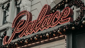 A neon sign displaying the word 'Palace' is prominently featured, surrounded by festive garlands with ornaments. The background consists of a classical building facade with decorative elements and pilasters.