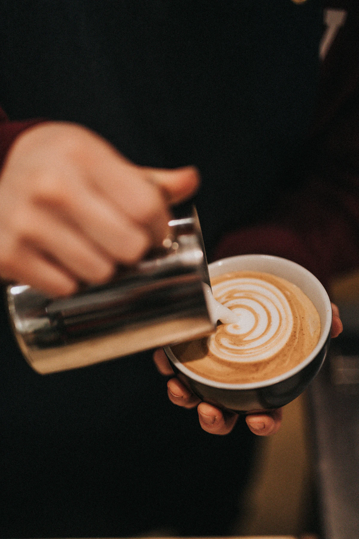 Barista pouring steamed milk from a stainless steel pitcher into a cup for latte art