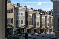 A row of modern townhouses with multiple floors and large windows, characterized by neutral colors and clean architectural lines. The houses are set closely together and feature small trees and landscaping in front. A clear blue sky with some clouds is visible above.