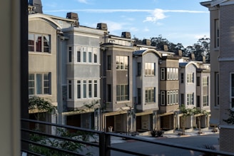 A row of modern townhouses with multiple floors and large windows, characterized by neutral colors and clean architectural lines. The houses are set closely together and feature small trees and landscaping in front. A clear blue sky with some clouds is visible above.