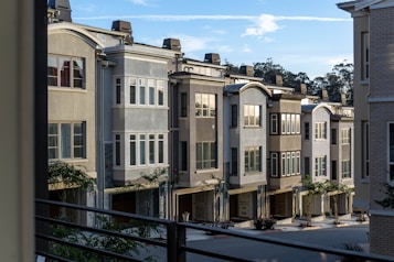 A row of modern townhouses with multiple floors and large windows, characterized by neutral colors and clean architectural lines. The houses are set closely together and feature small trees and landscaping in front. A clear blue sky with some clouds is visible above.