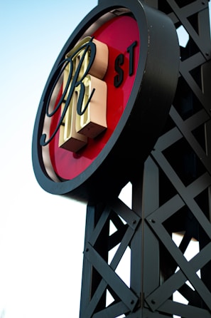 A circular sign with large, stylized letters in the center placed on a lattice-like metal framework. The background of the sign is red, with letters 'RH' prominently displayed. The letters 'ST' appear smaller on the side. The structure has a modern industrial design.