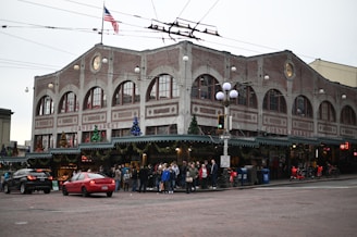 A bustling corner market building with arched windows and festive decorations including Christmas trees and lights. A group of people gather on the sidewalk, waiting at the corner while cars pass by on a cobblestone street. An American flag is visible atop the building.
