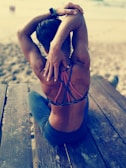 A person is sitting on a wooden bench at the beach, stretching their arms above their head. They are wearing a sporty outfit with a detailed crisscross back design. The beach setting is blurred in the background.
