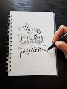 A hand is holding a pen and writing on a dotted notebook page. The handwritten text reads 'Always start your day with a cup of Positivitea' in elegant, decorative script. The notebook has a spiral binding on the left side, and the background is a dark, wooden surface.