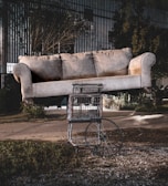 A large beige sofa is precariously balanced on top of a shopping cart with a single wheel, set outdoors. The background features a street, overgrown grass, and some industrial buildings, giving a surreal and somewhat abandoned atmosphere.