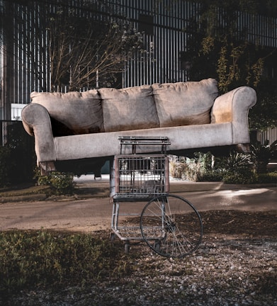 A large beige sofa is precariously balanced on top of a shopping cart with a single wheel, set outdoors. The background features a street, overgrown grass, and some industrial buildings, giving a surreal and somewhat abandoned atmosphere.