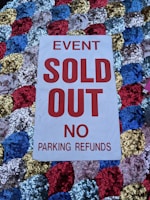 A white sign with bold red text stating 'EVENT SOLD OUT NO PARKING REFUNDS' is placed on a colorful sequined surface composed of blue, red, gold, and silver circular patterns.