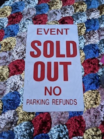 A white sign with bold red text stating 'EVENT SOLD OUT NO PARKING REFUNDS' is placed on a colorful sequined surface composed of blue, red, gold, and silver circular patterns.